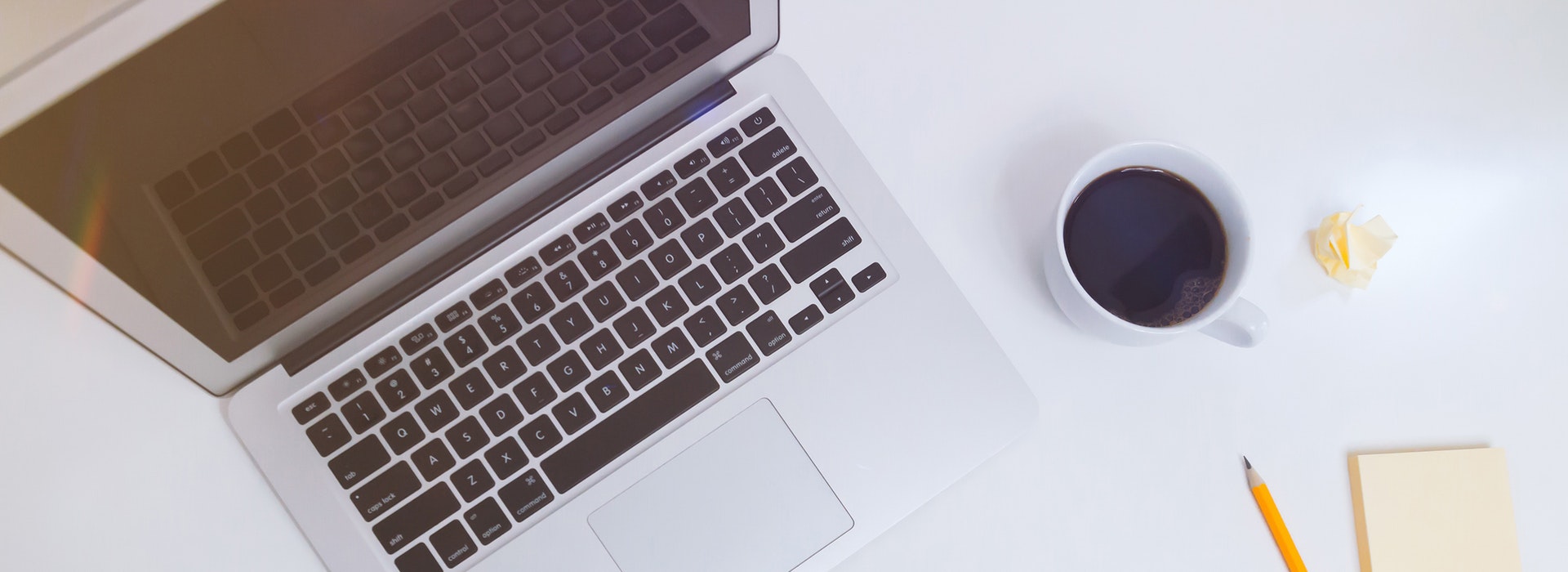 A black and silver laptop keyboard sits beside a cup of black coffee and a sharpened pencil