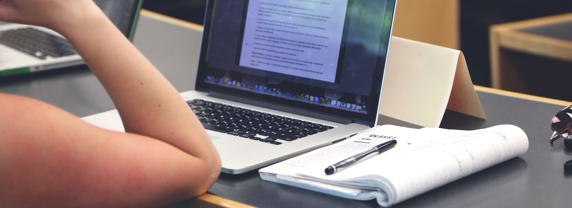 Close up of a person's arm resting on a desk with a laptop and notebook