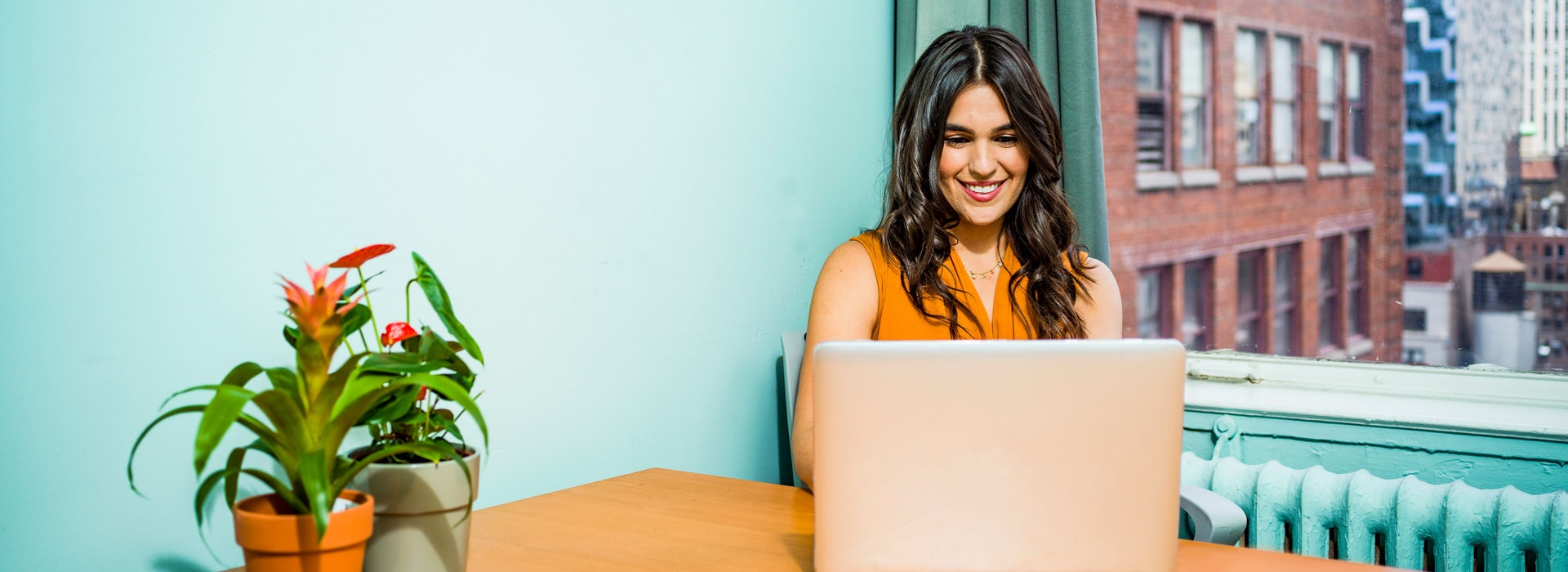 A woman with long dark hair sits against a turquoise wall, smiling at a laptop