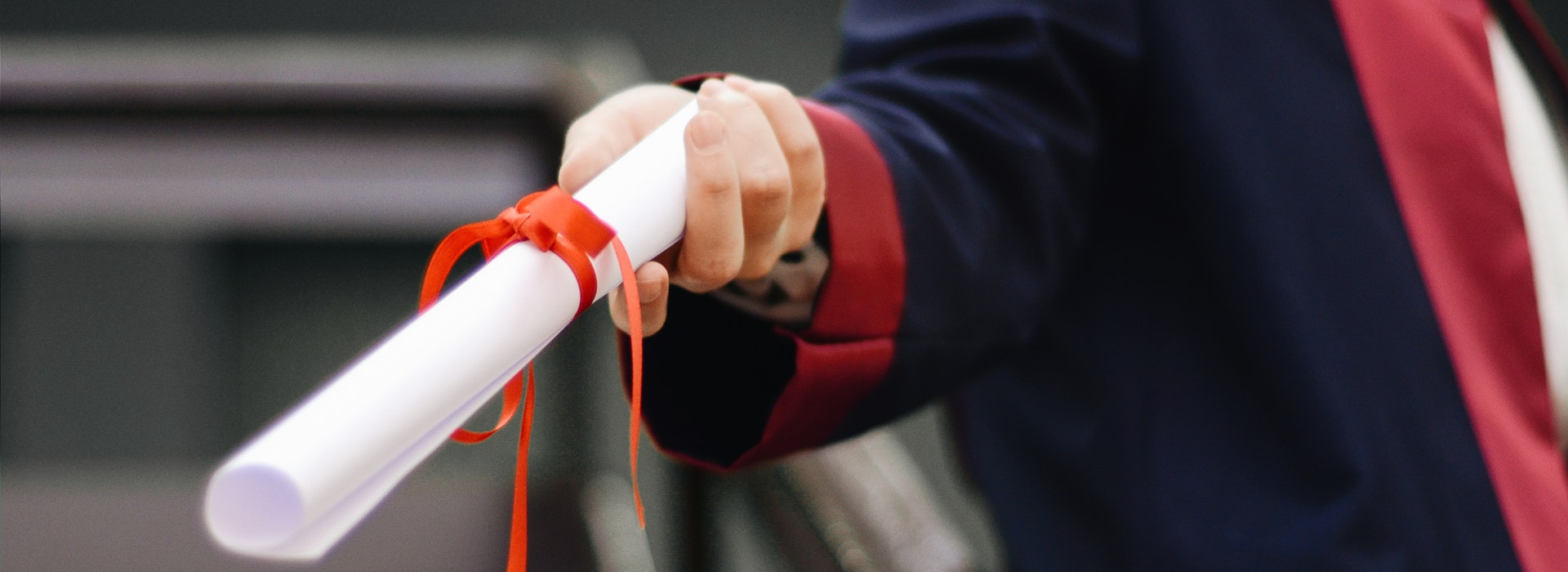 Someone’s hand holds a white, rolled diploma tied with a red ribbon