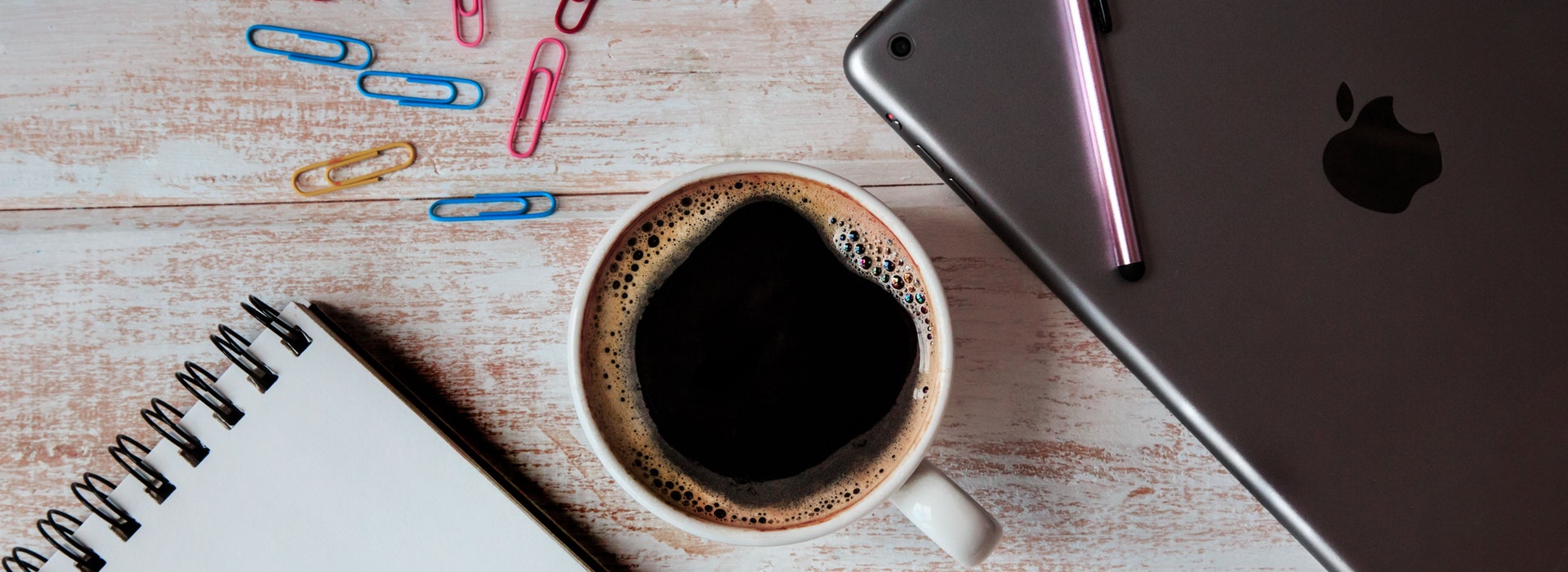 A silver laptop, colorful paper clips and a cup of coffee are arranged on a white desk