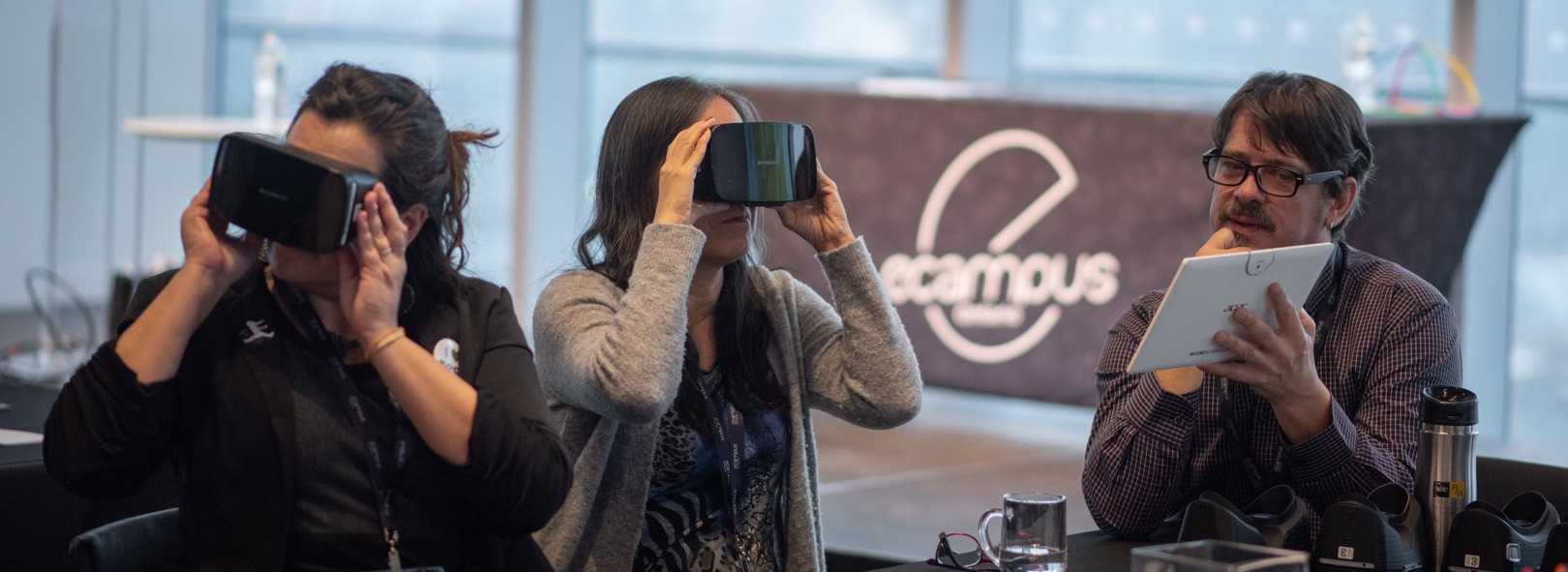 Two women sit at a table with virtual reality headsets, while a man with a notebook looks on