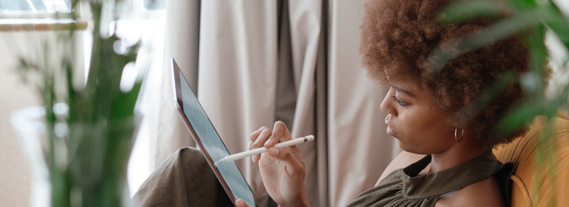 A young woman sits among green plants reading a tablet