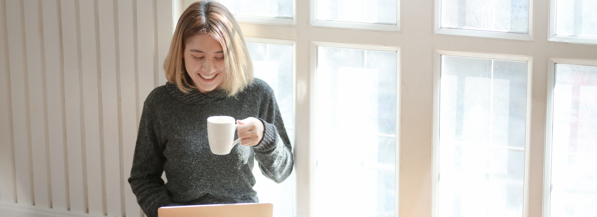 A woman sits in a window, drinking coffee and typing on a laptop