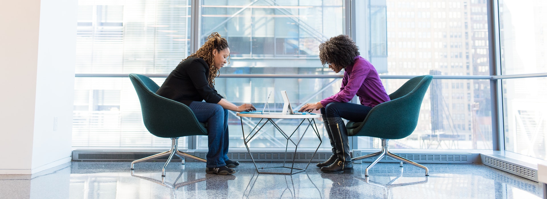 Two women sit across from each other typing on laptops