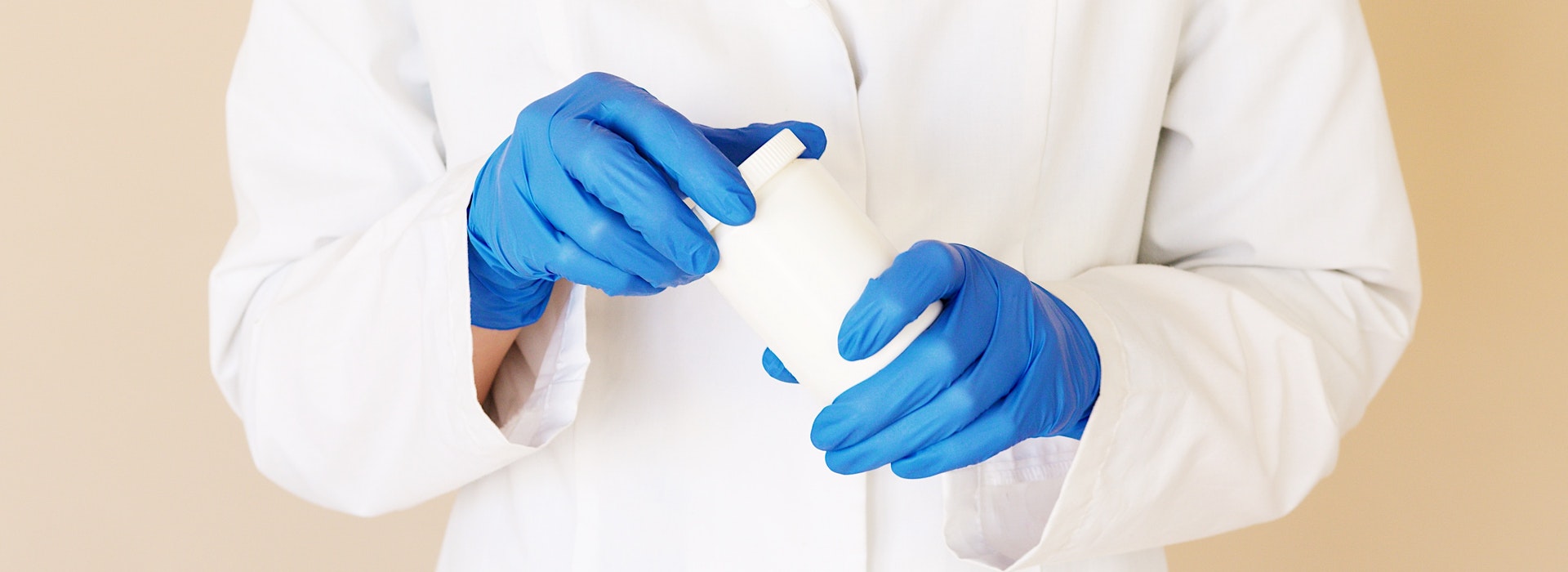 Photo of a person wearing lab coat and blue medical gloves holding a plastic bottle