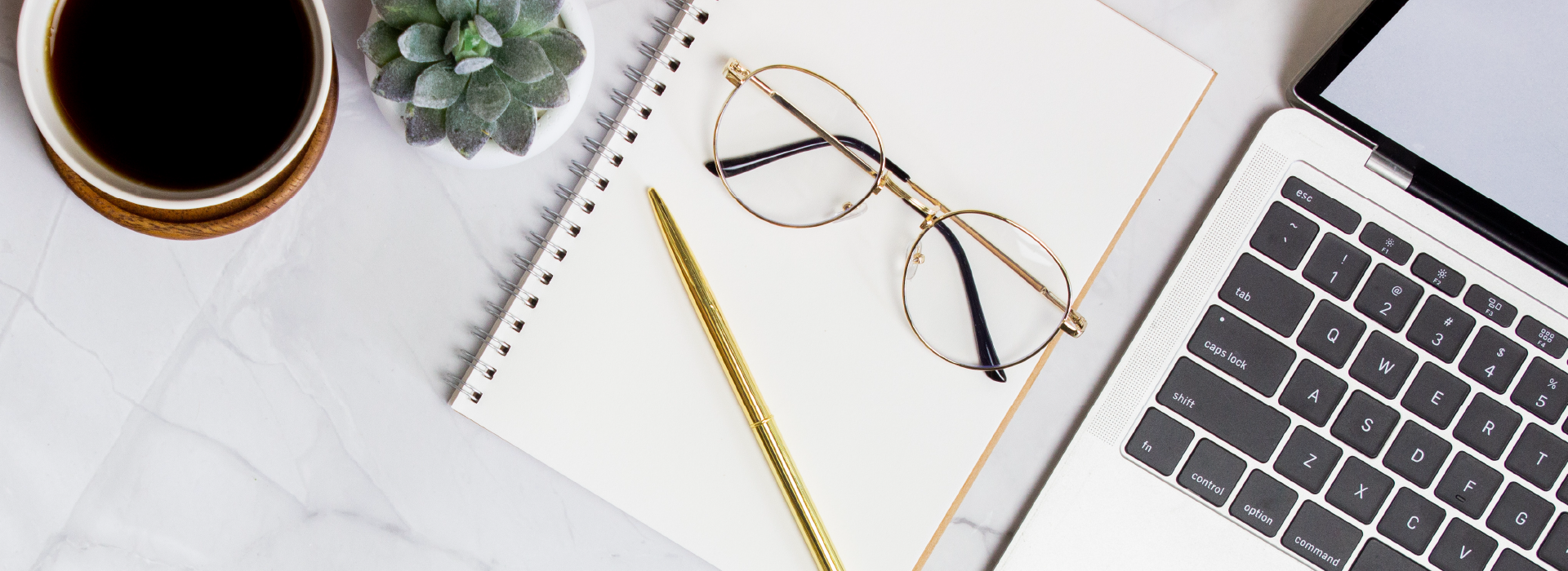 A notebook, pair of glasses, a cup of coffee and an open laptop on a marble table